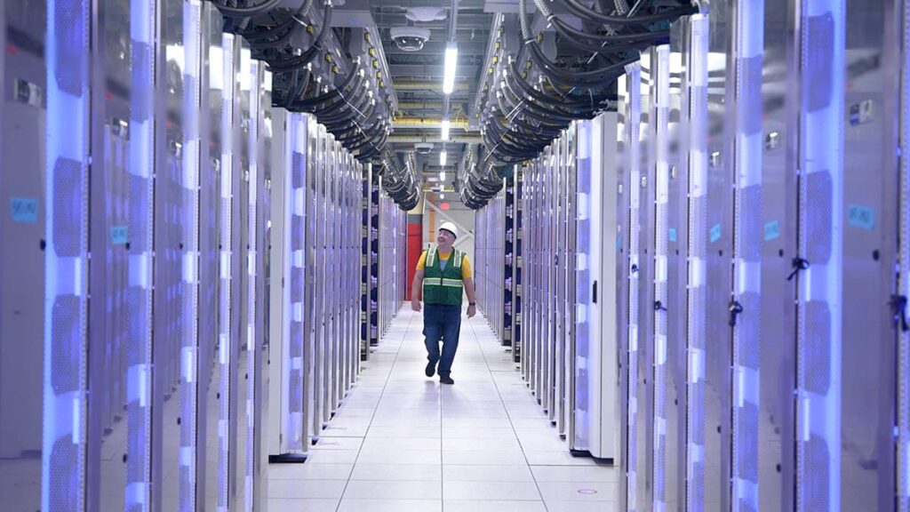 A view of the interior of Elon Musk / X's new AI server farm. Ranks of server banks retreat into the distance. A workier in a hard hat walks the corridor between them.